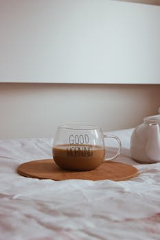 A warm cup of coffee in a transparent glass mug on a cozy bed setting perfect for a relaxing morning.