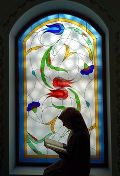 A woman reading a book in prayer near a stained glass window in a Turkish mosque.