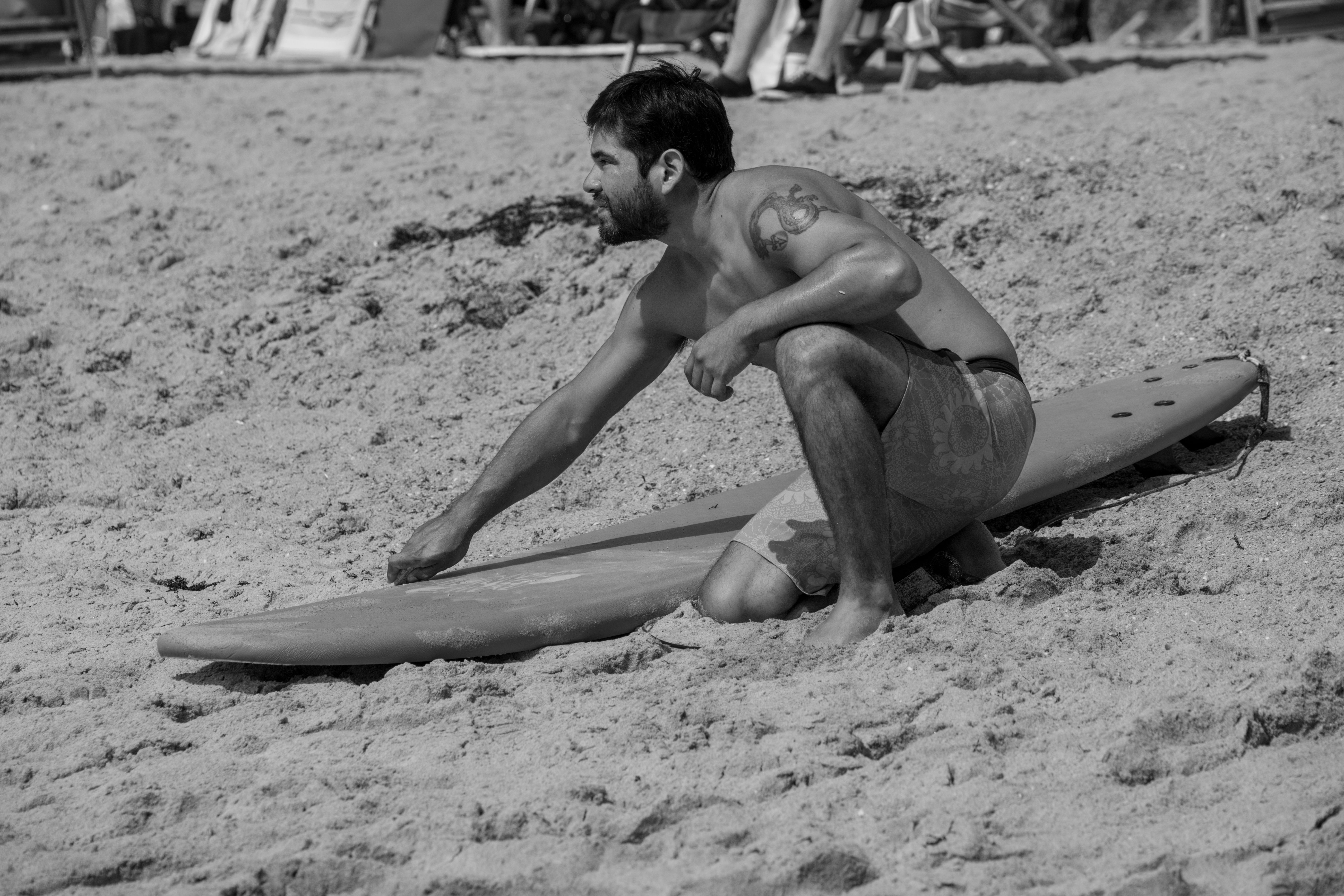 Grayscale photo of a tattooed surfer adjusting his board on the sandy Cocoa Beach.