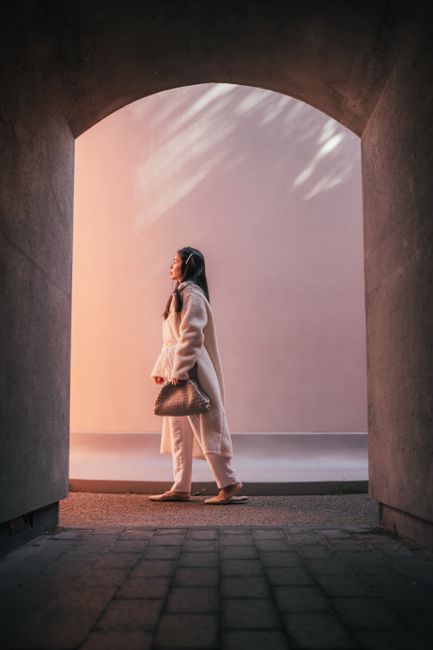 Elegant woman with long hair posing under an archway during sunset, dressed in modern fashion.