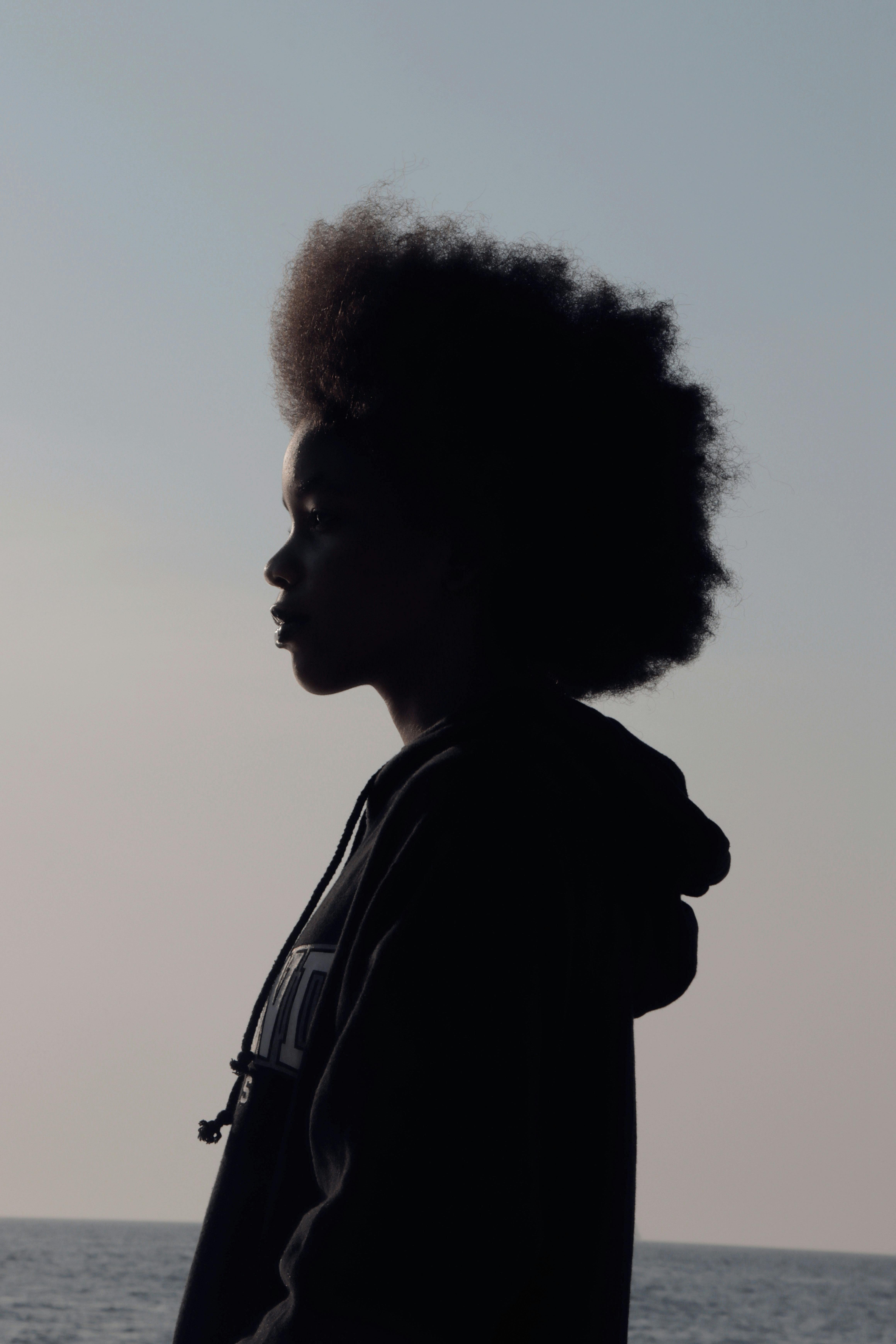 A serene silhouette of a child with afro hair gazing at the sea horizon during sunset.