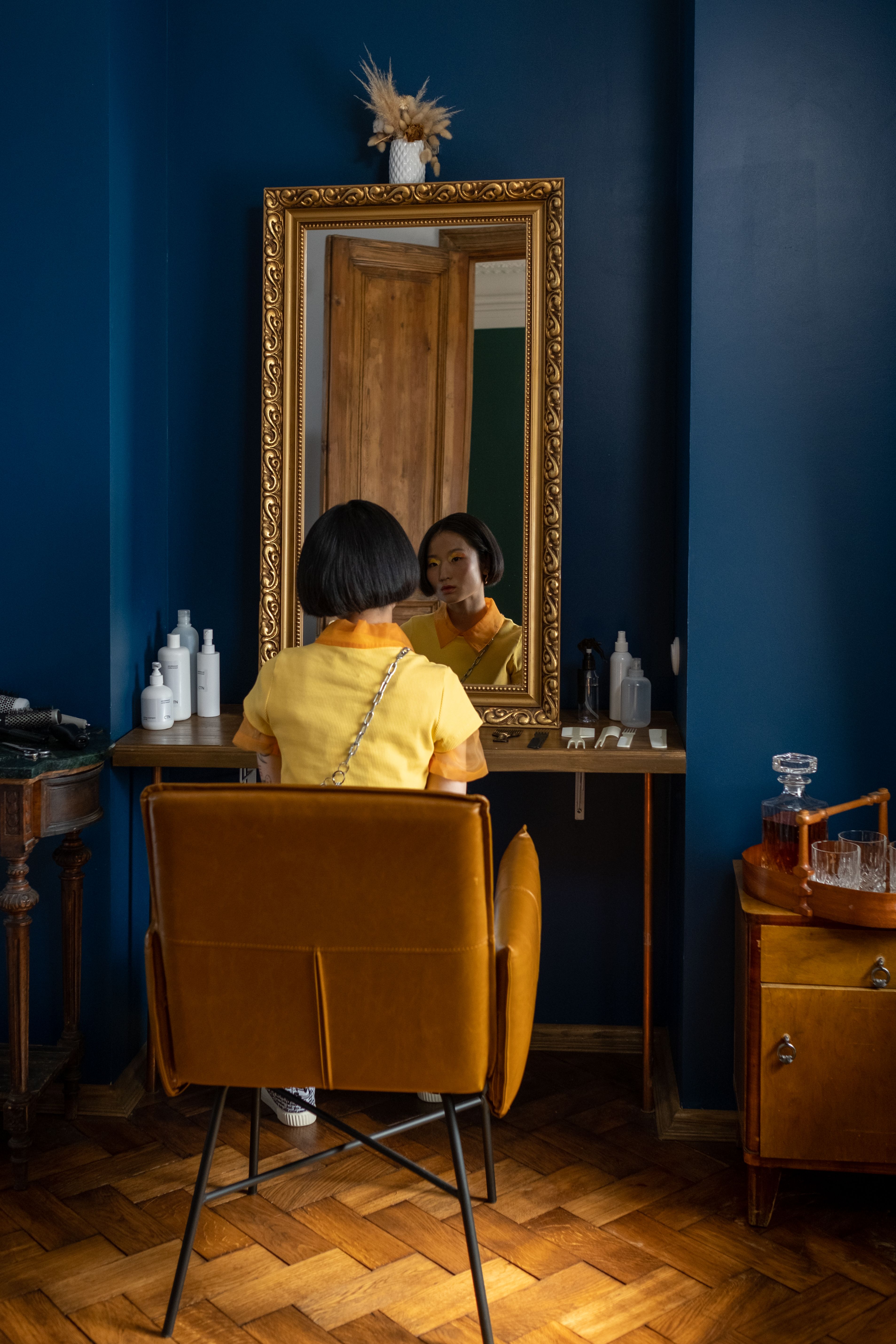 A woman with short black hair in a yellow outfit seated at a vanity mirror stylish room.