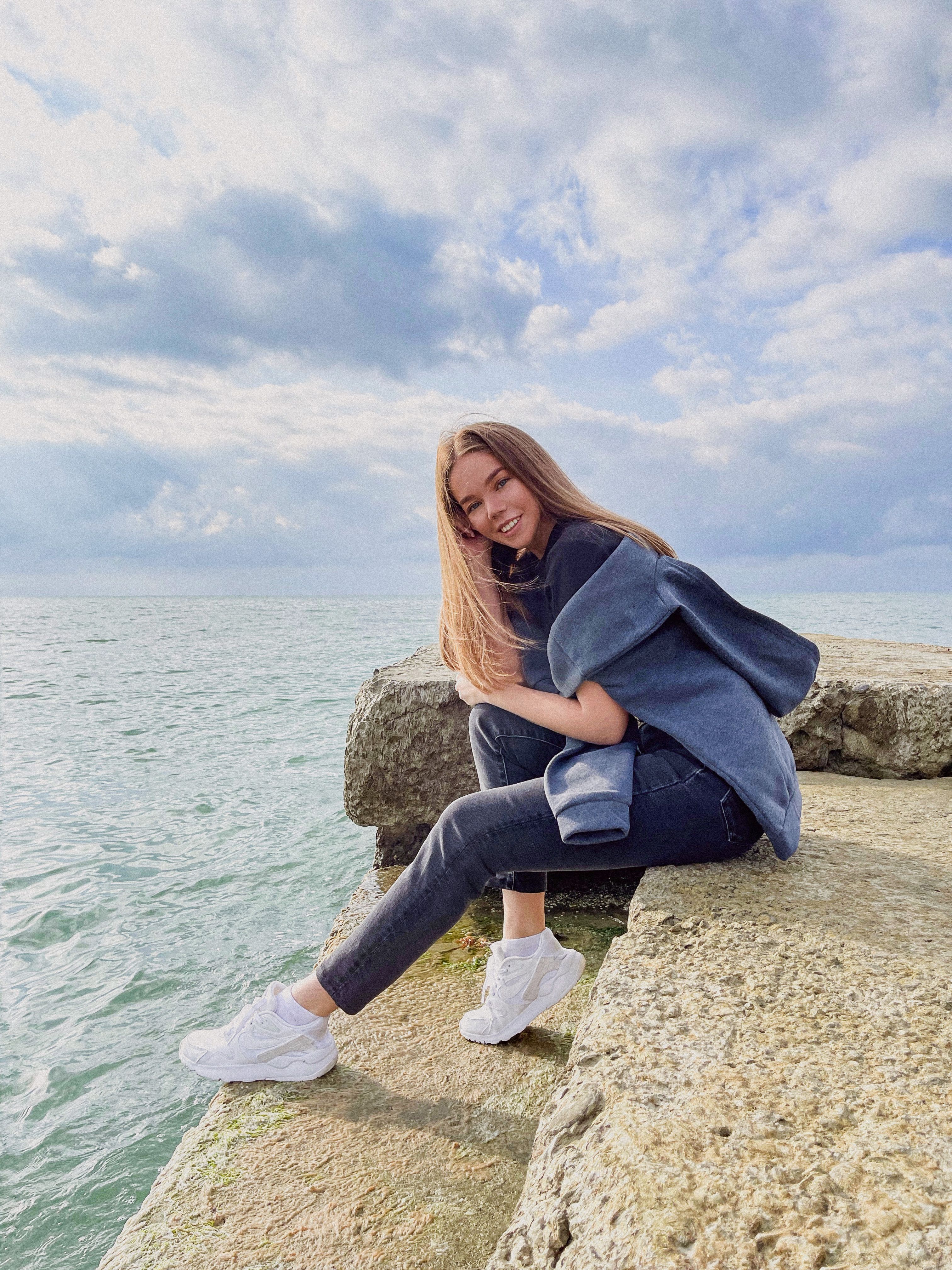 Smiling woman sitting on a rocky shore with a vast sea backdrop under a cloudy sky.