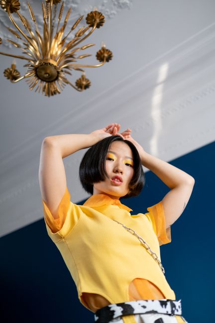 Stylish woman in yellow outfit with creative makeup poses indoors beneath ornate chandelier.