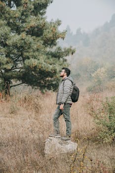 A man with a backpack stands on a rock in a misty forest during autumn, surrounded by trees.