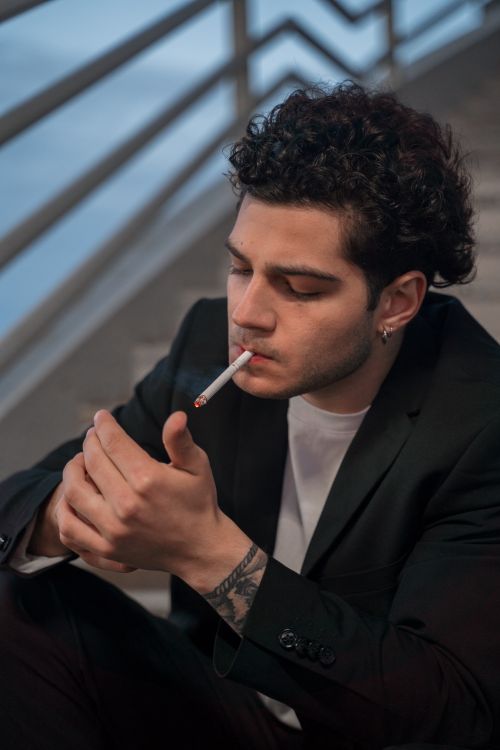 Free Young man with curly hair smoking on outdoor stairs, lost in thought. Stock Photo