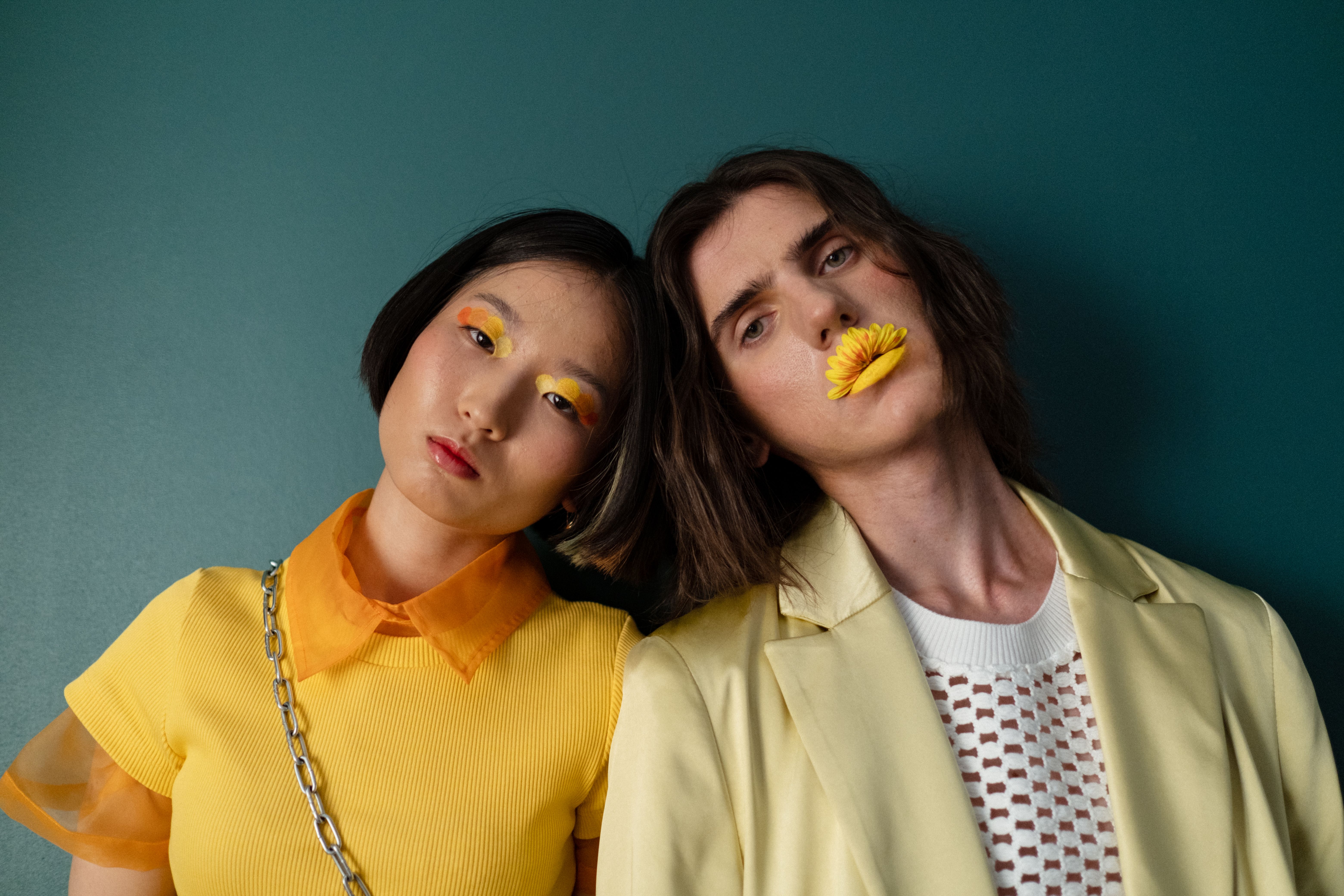 Two individuals in stylish yellow outfits and creative makeup pose against a green background.