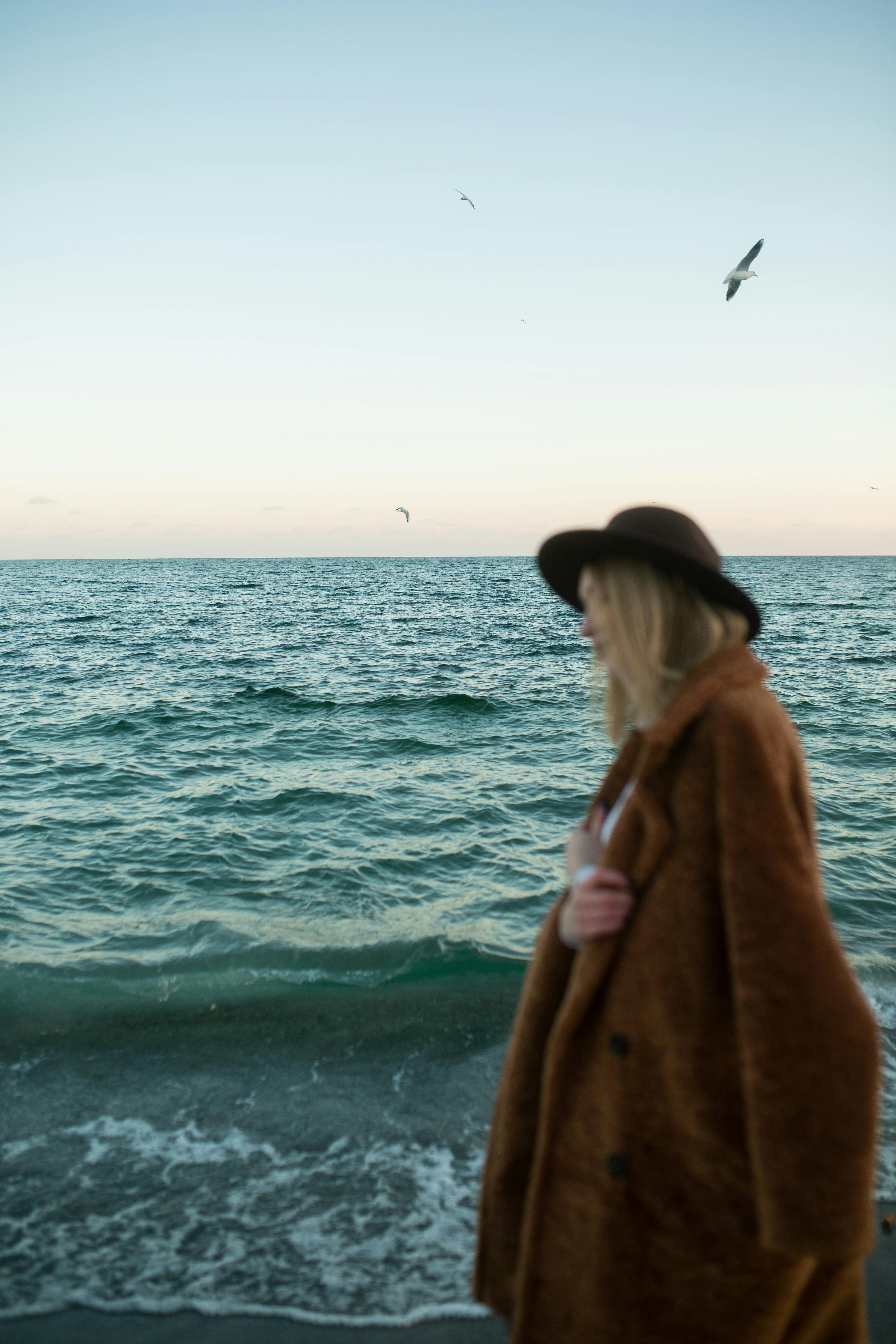 A woman in a brown coat and hat enjoys the seaside view at sunset, with waves and birds in the background.
