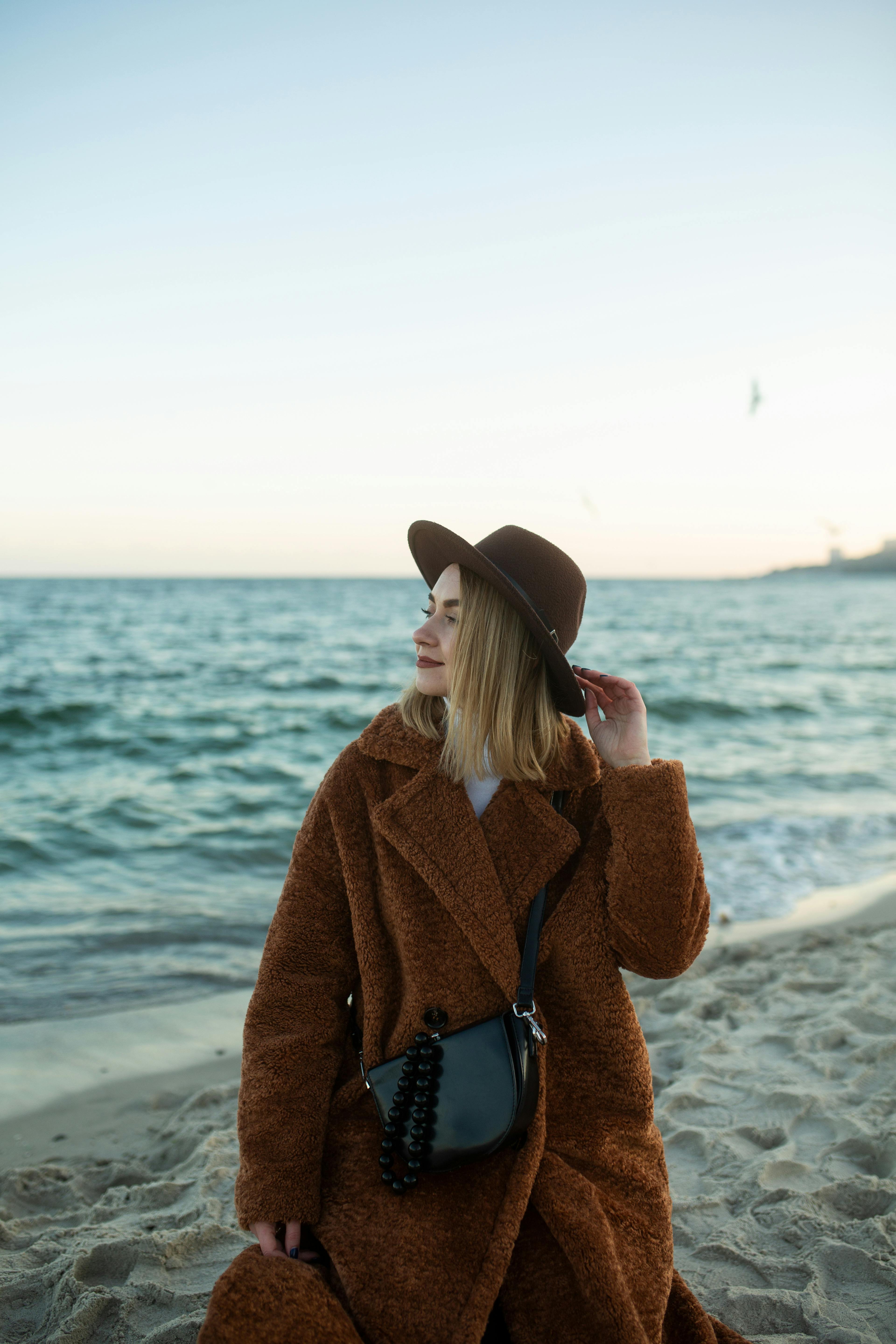 A stylish woman sits at the seashore, wearing a brown coat and hat, enjoying the ocean view.