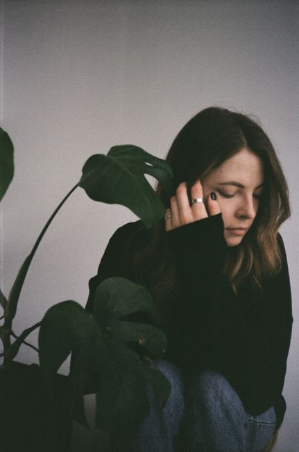 Caucasian woman sitting indoors with eyes closed, surrounded by monstera plant leaves.