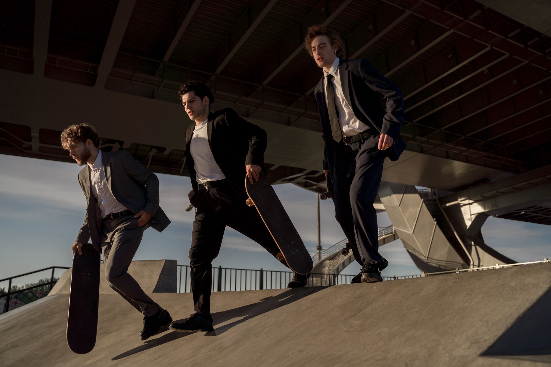 Three men in suits skateboarding in a modern urban skate park under a bridge.