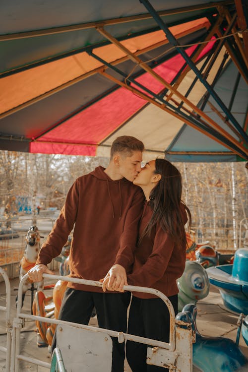 A Couple Wearing Maroon Hoodie Kissing Each Other while Standing Near an Amusement Ride