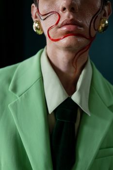 A close-up portrait of a person wearing a green suit with artistic red makeup and gold earrings.