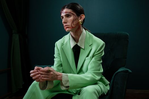 A person sits in a green suit with artistic makeup and earrings in a dark room.