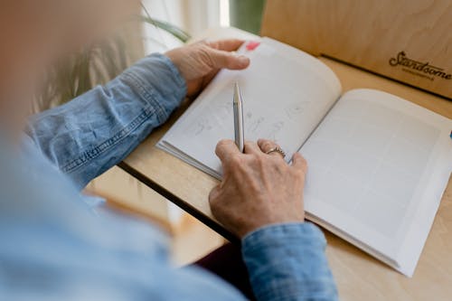 Person in Blue Denim Jacket Holding Pen Writing on White Paper