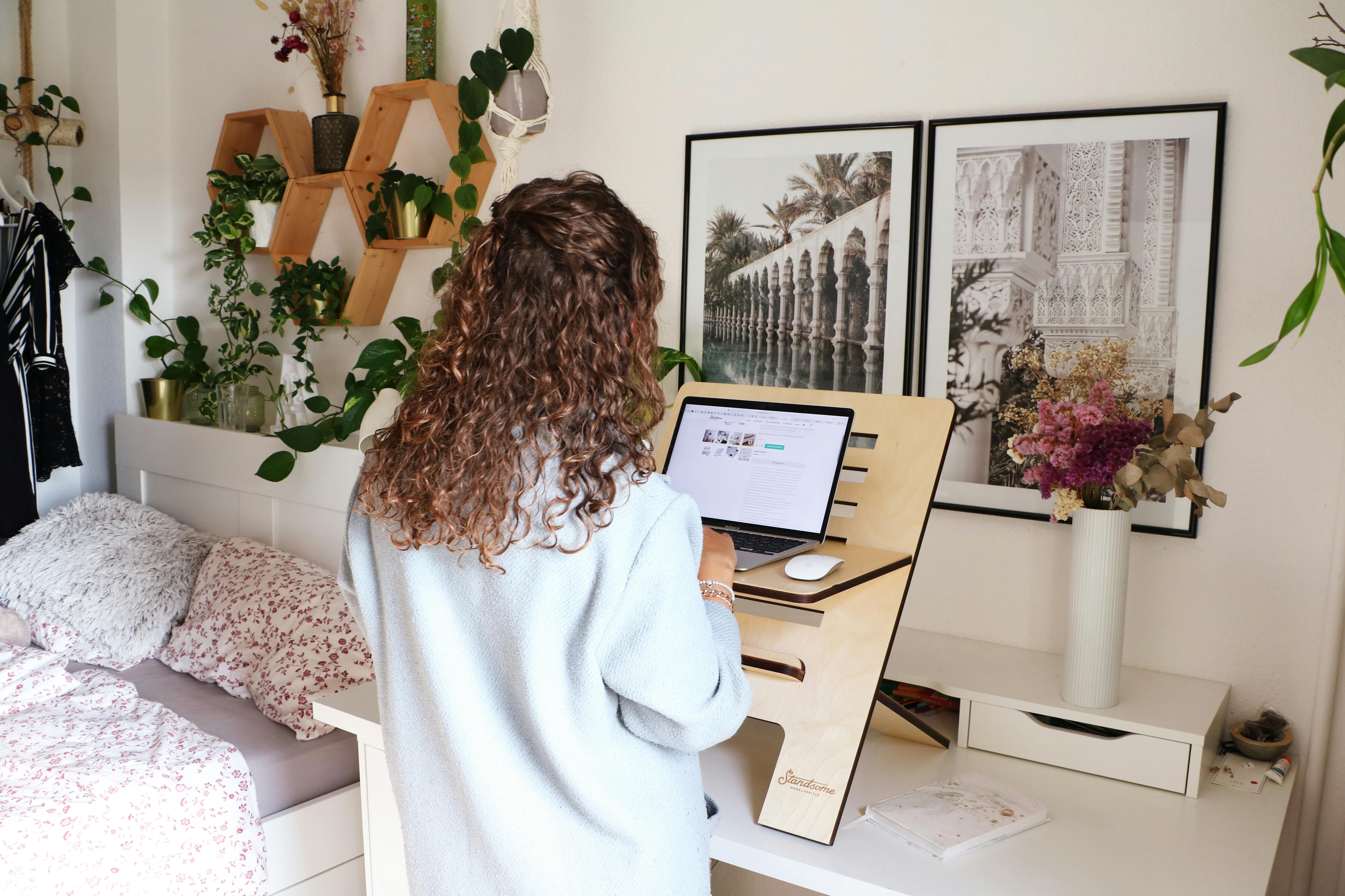 Person using a compact standing desk in a clutter-free bedroom with laptop and papers