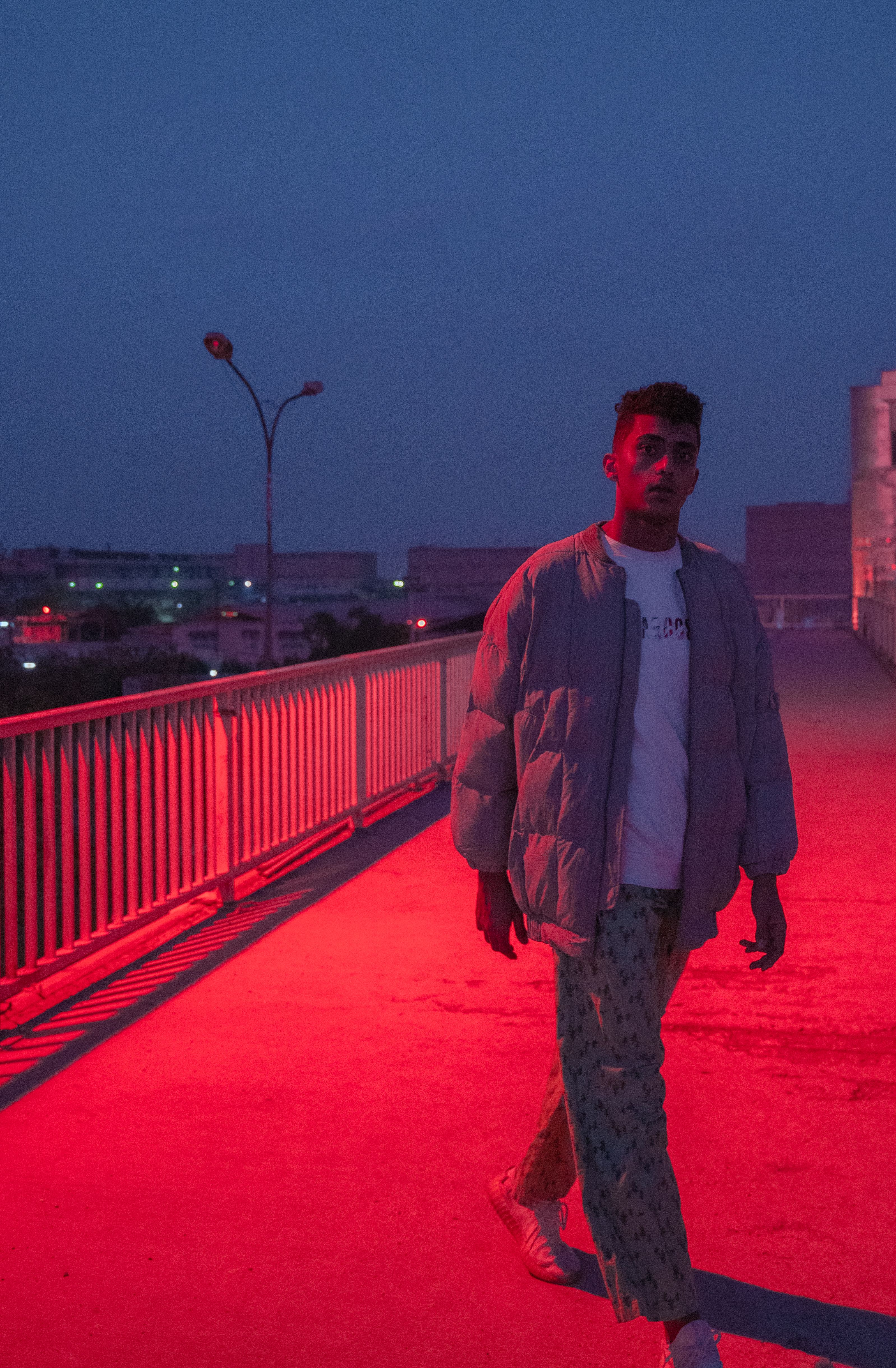 Stylish man in casual attire walking on a bridge lit by red lights during twilight.