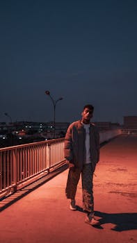 A man in casual clothes walking on a rooftop at nightfall, next to a metal railing.