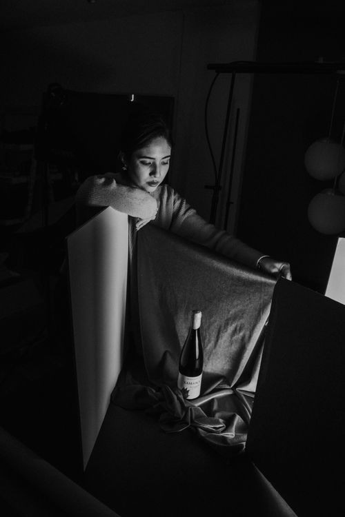 Free Black and white image of a woman setting up a wine bottle display with soft lighting in a studio. Stock Photo