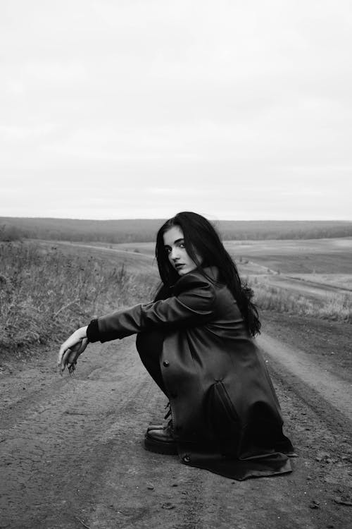 Grayscale Photo of Woman in Black Long Sleeve Dress Sitting on Dirt Road