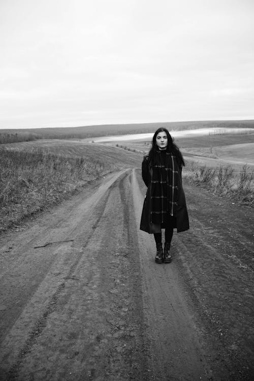 Black and White Photo of a Woman in a Coat Standing on a Dirt Road