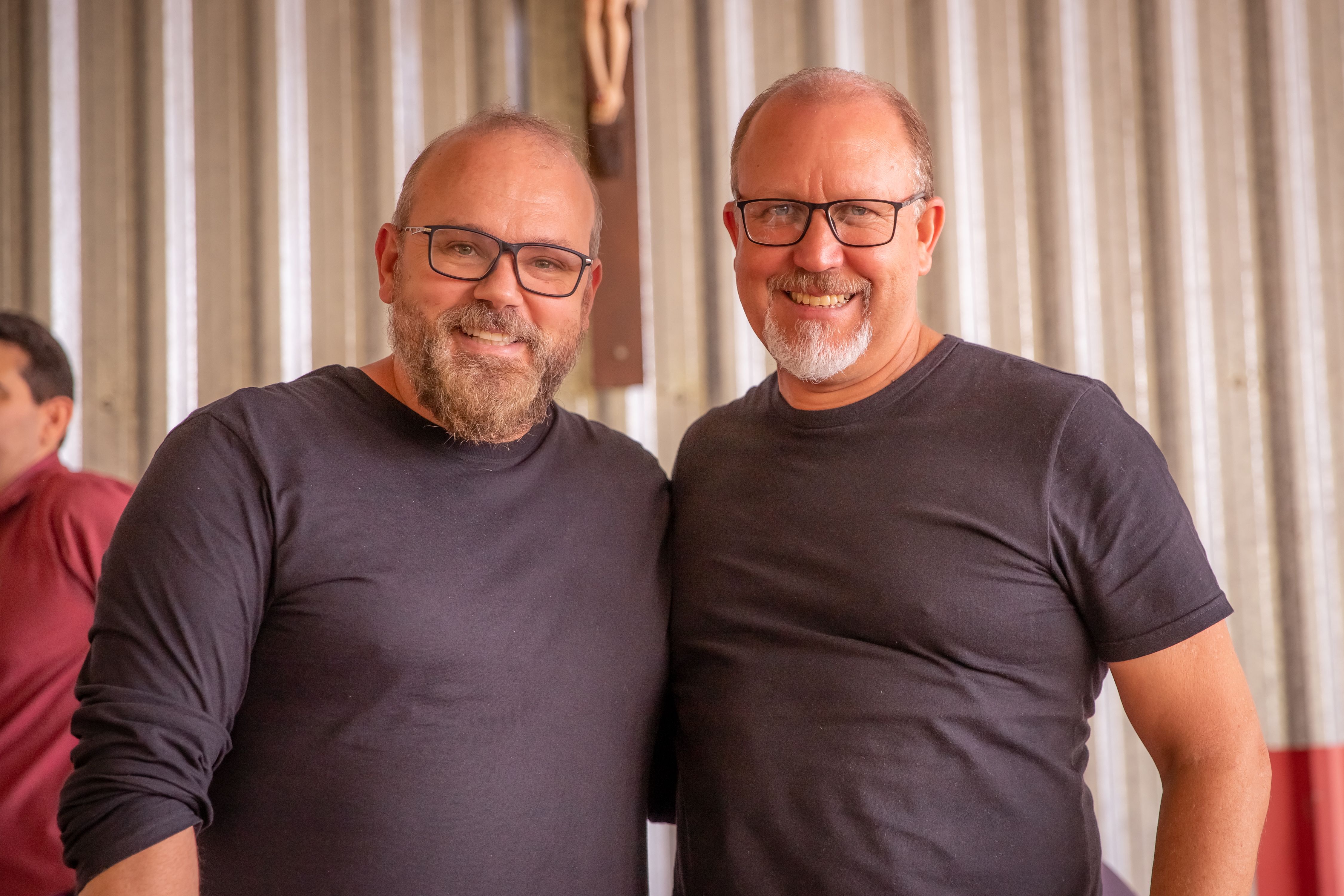 Two smiling men with eyeglasses and facial hair pose happily indoors, showcasing friendship.