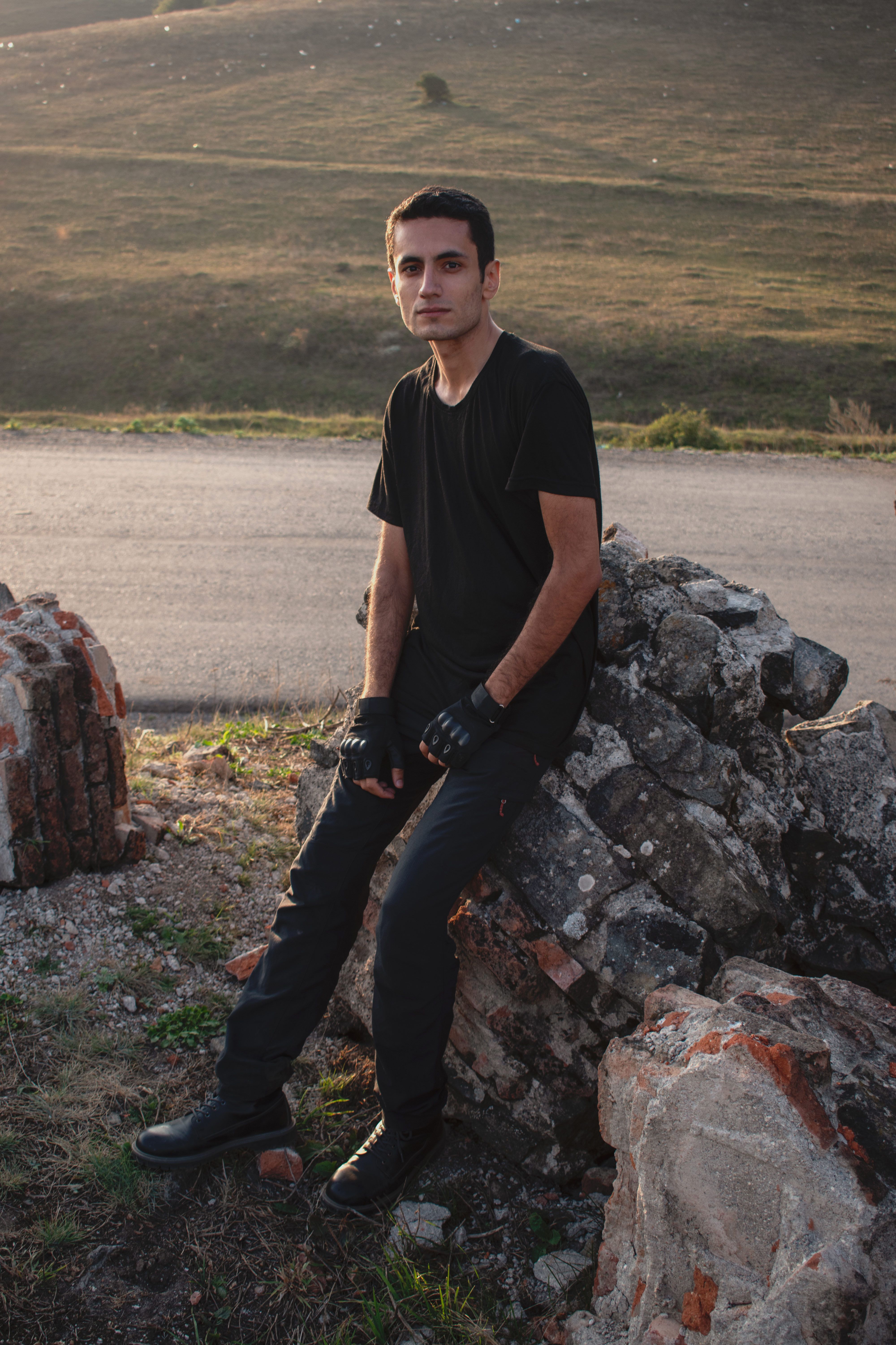 A man in a black outfit poses on rocks in a serene outdoor setting.
