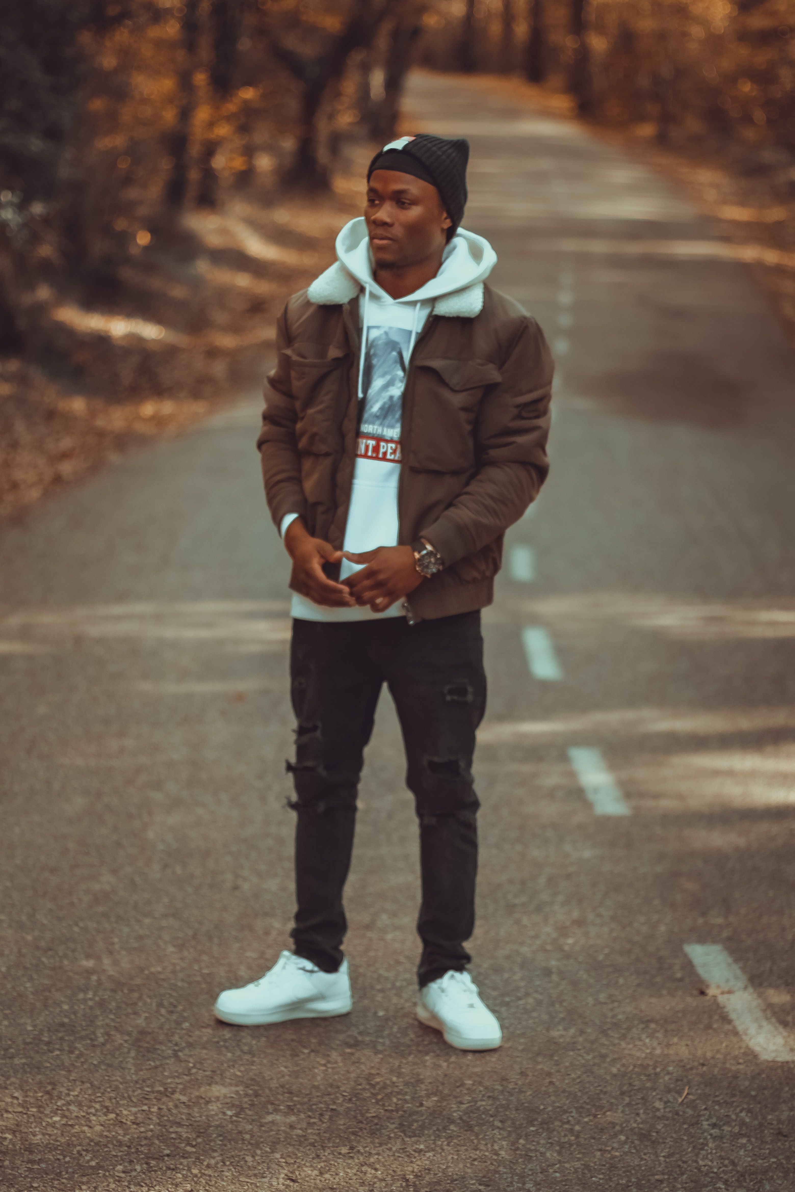 A fashionable man poses confidently on a tree-lined road during autumn, showcasing modern street style.