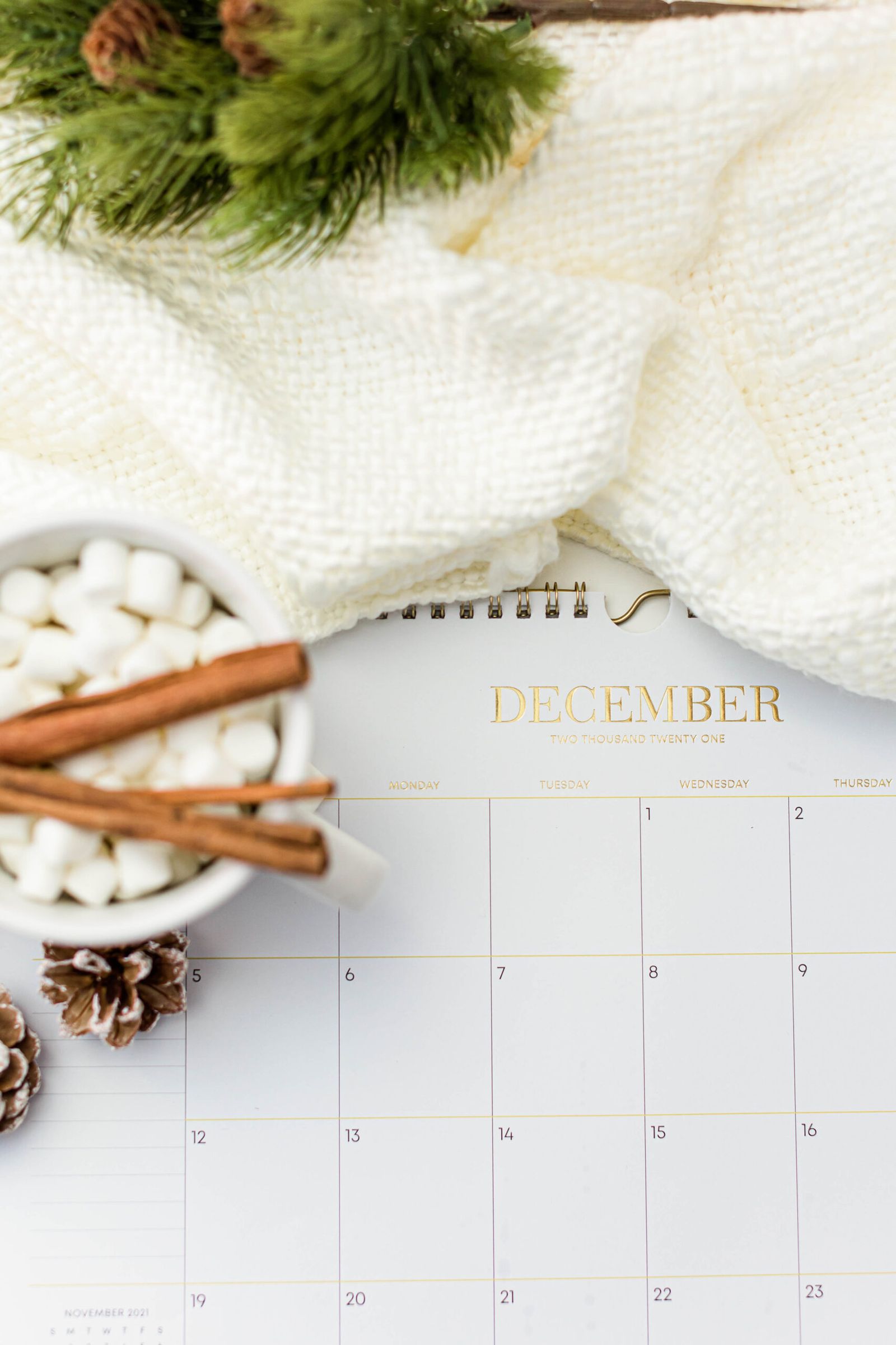 December calendar beside hot cocoa, evergreen branches, cinnamon sticks, and a soft white blanket.