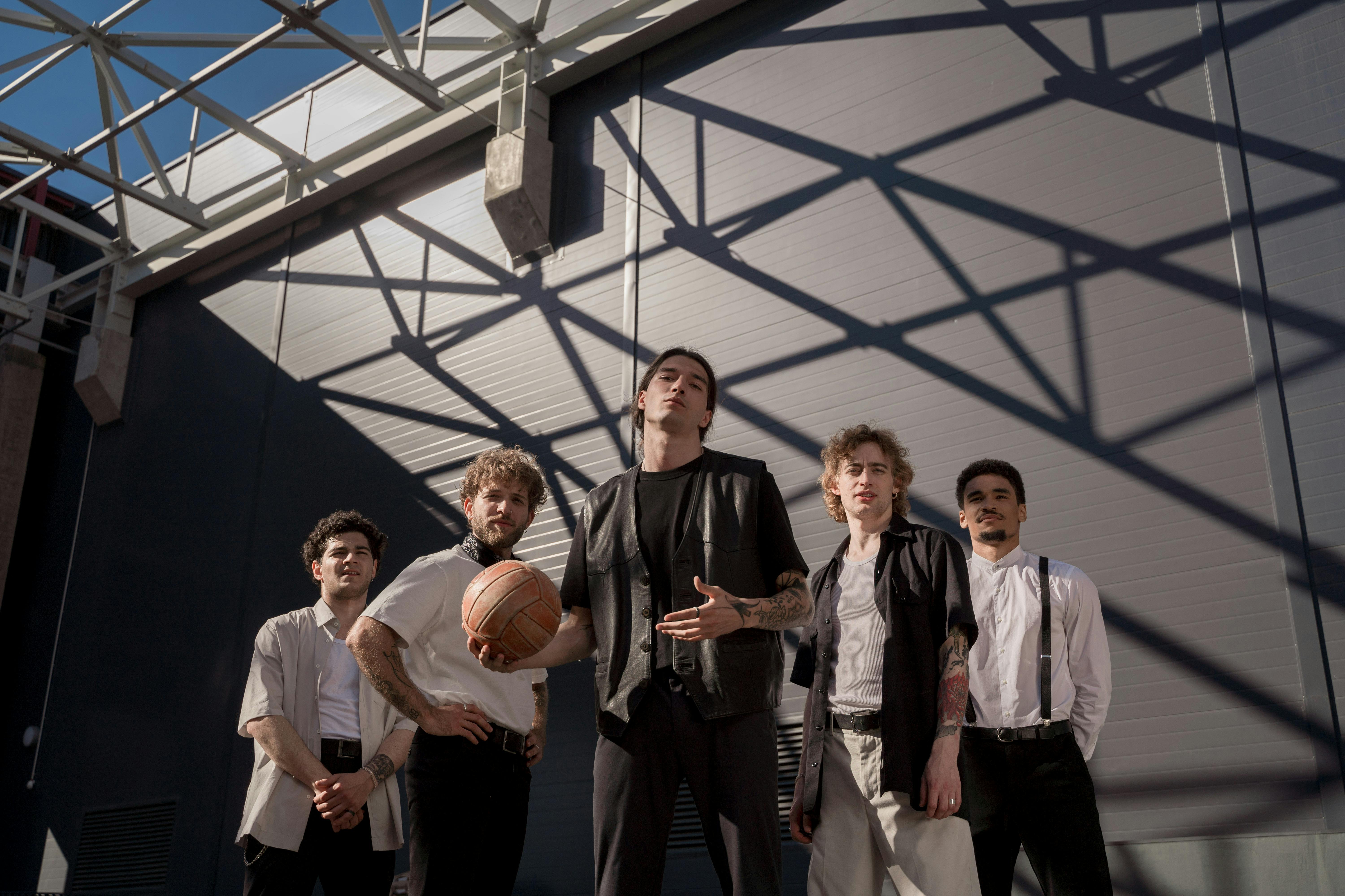 Five stylish men pose outside with a basketball, shadows creating a dynamic city backdrop.