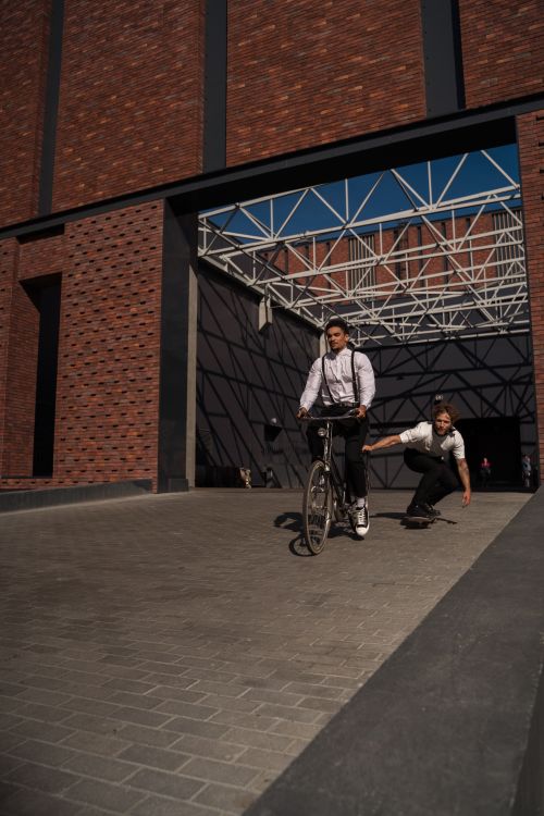 Free Two young adults biking and skateboarding outside a modern brick building, enjoying a sunny day. Stock Photo