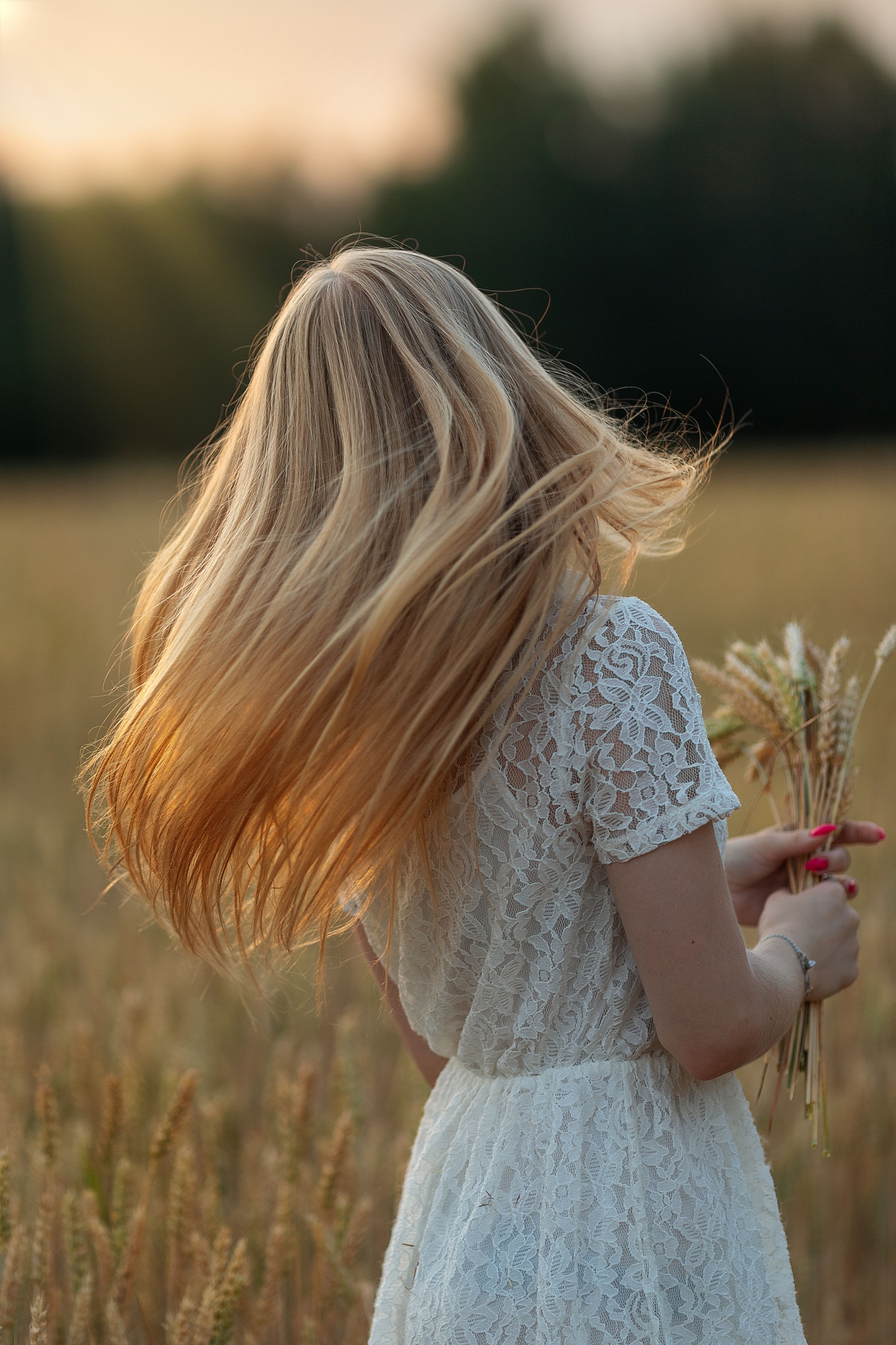 A woman with flowing blonde hair in a white lace dress stands in a sunlit wheat field holding wheat stalks.