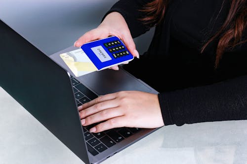 A Person in Black Long Sleeves Holding a POS Device with Card while Typing on a Laptop