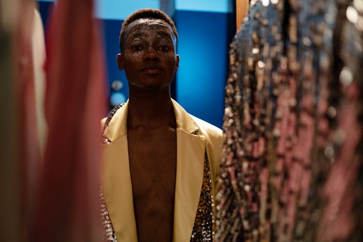 Young Man In Stylish Outfit Standing Behind Hanging Clothes