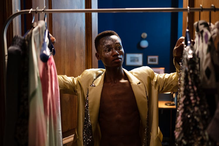 Man In Glitter Makeup Looking Through Clothes Hanging In Wardrobe