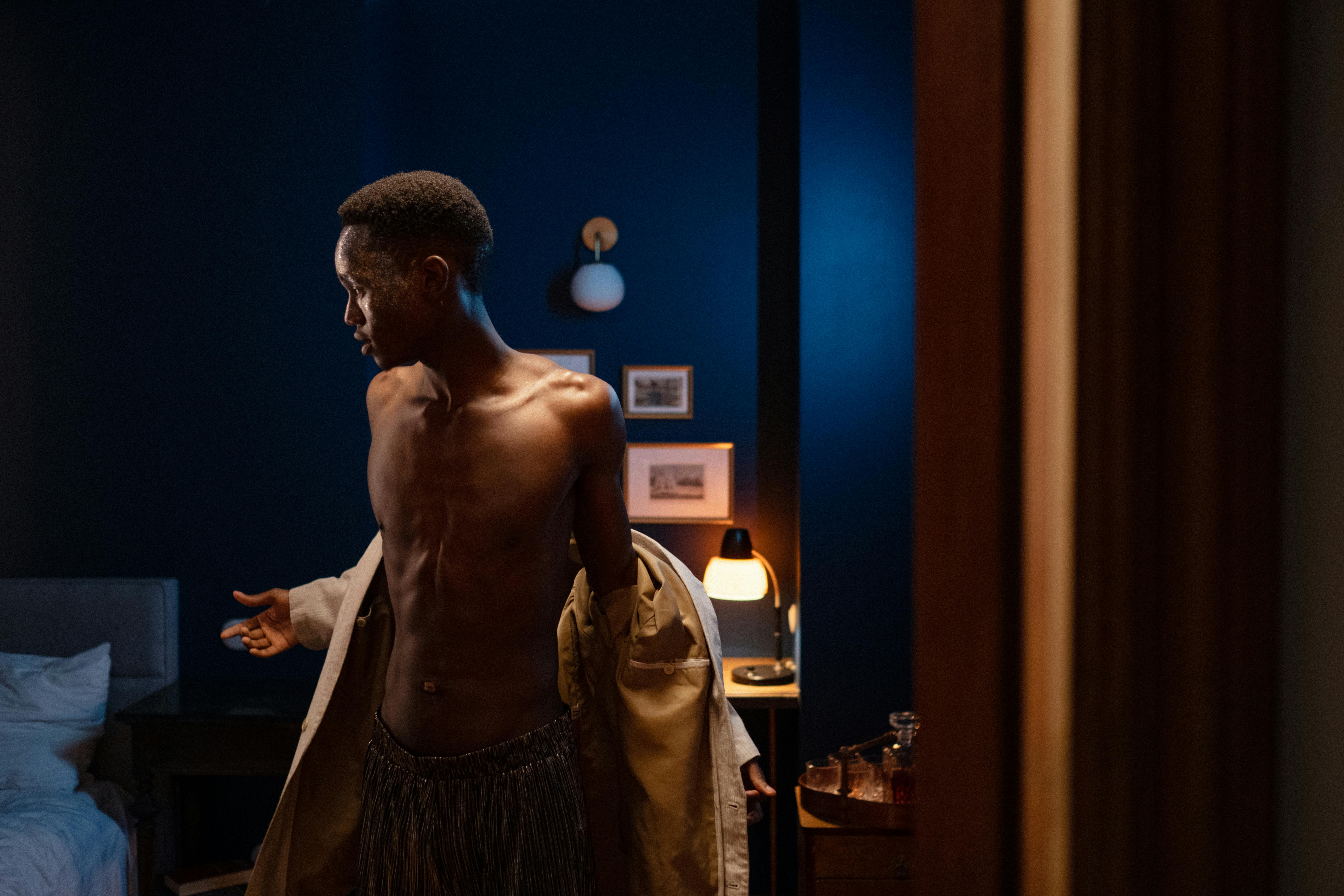 young man with naked torso standing in dark bedroom and dressing up