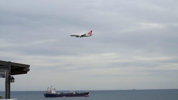 Airplane approaching over the sea with a cargo ship below under a cloudy sky.