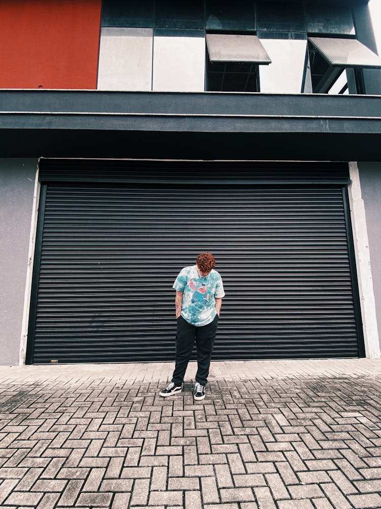 A Man In In Blue Shirt And Black Pants Standing Near Steel Rolling Shutter While Looking Down