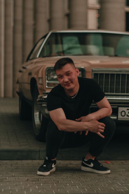Free Asian man poses in casual black attire squatting beside a vintage car on an urban street. Stock Photo