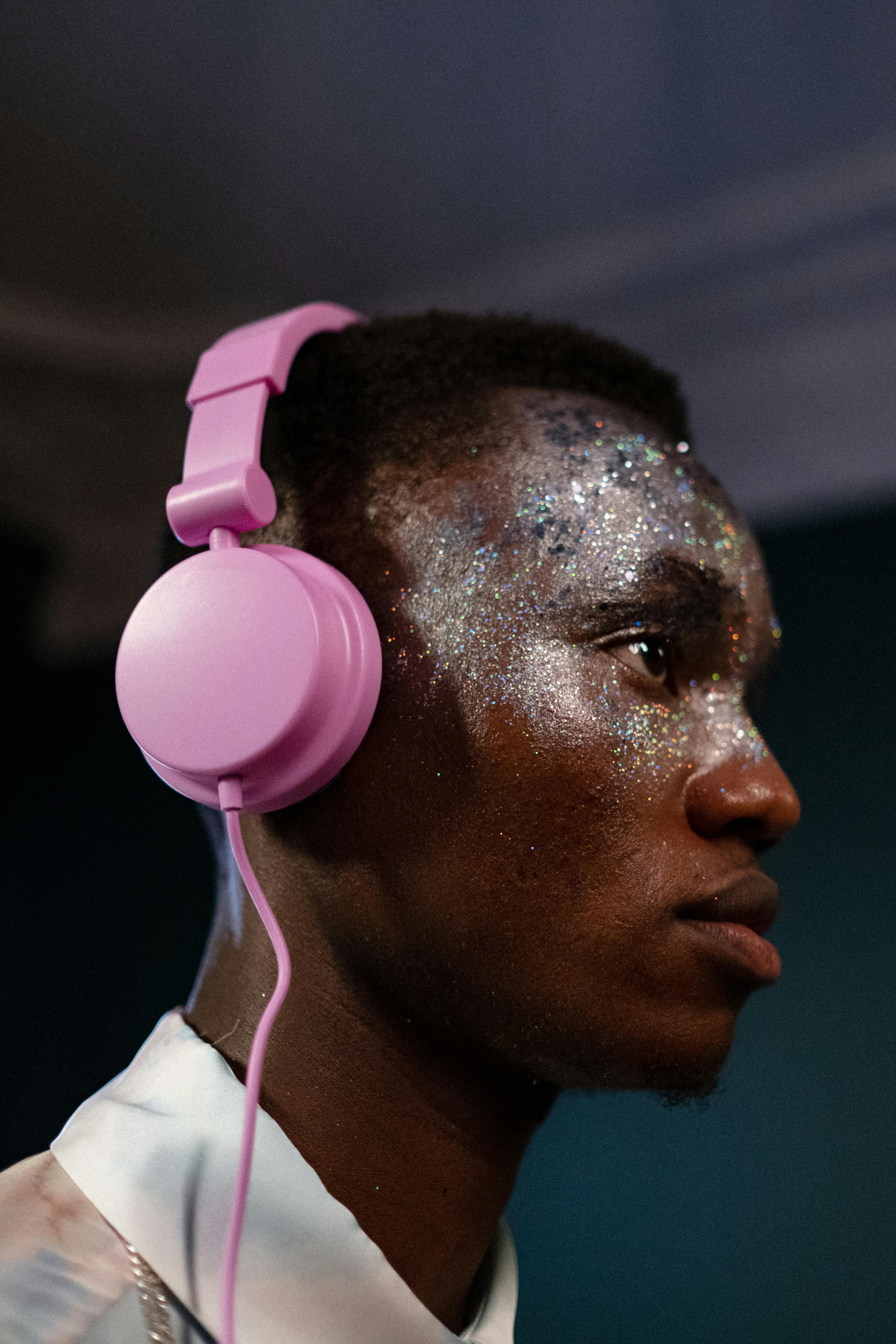 Portrait of a young man with glitter makeup wearing pink headphones indoors.