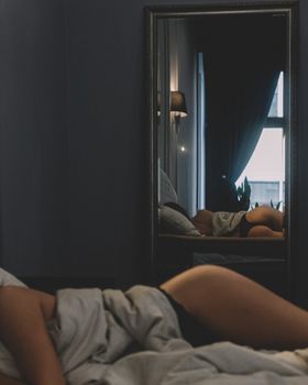 A woman is lying in bed with a focus on her reflection in a bedroom mirror, creating a serene ambiance.