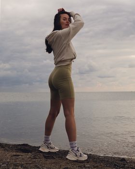 Woman in casual wear standing on a beach shore, posing with a cloudy sky backdrop.