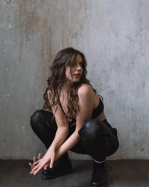 A Woman in Black Brassiere and Black Pants Sitting Near Gray Wall while Looking Afar