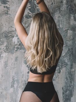 Back view of a blonde woman in black lingerie posing against a textured wall.