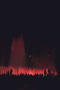 人们观赏着戏剧性的夜间喷泉表演，剪影映衬着充满活力的灯光效果