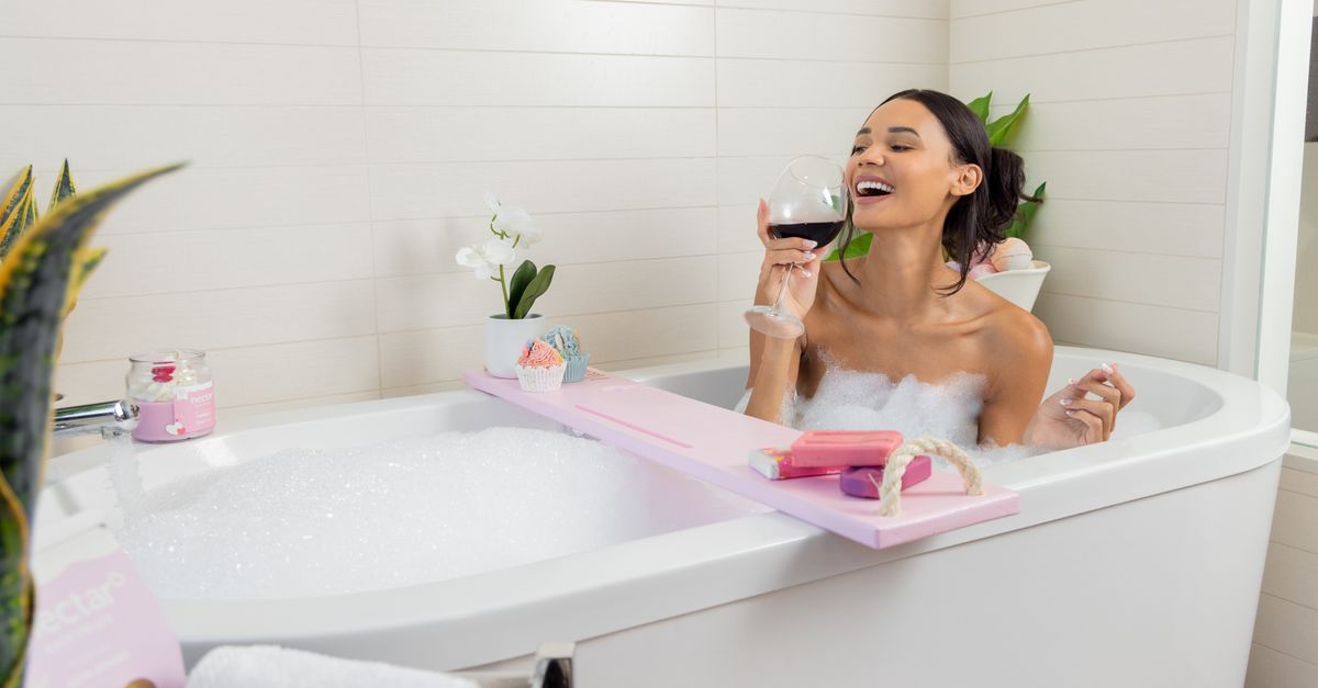 Woman enjoying a bubble bath with wine, surrounded by spa essentials in a modern bathroom.