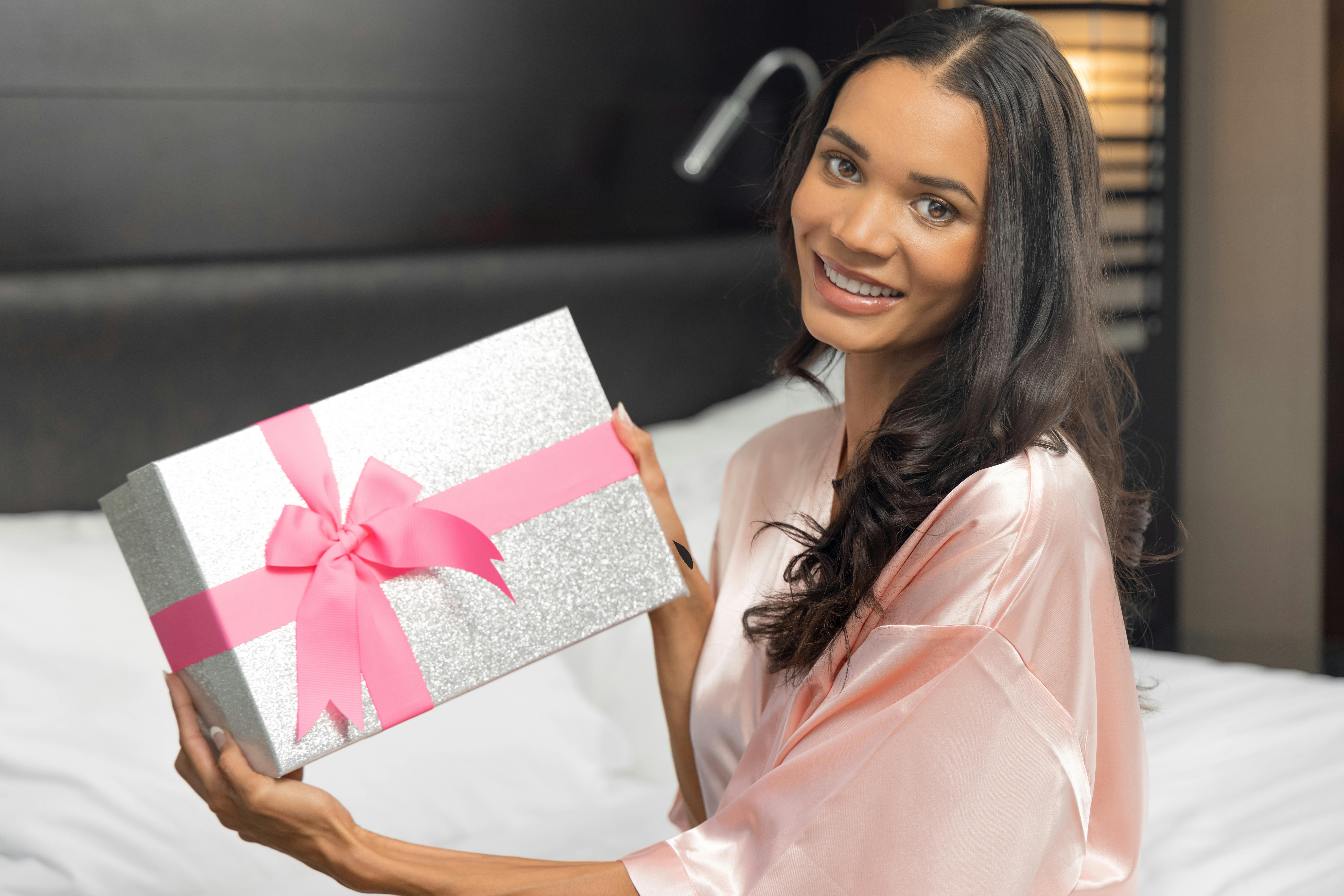 Happy woman in pink satin robe holding a gift box with pink ribbon, sitting indoors.