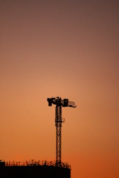 A tower crane silhouetted against a colorful sunset sky at a construction site.