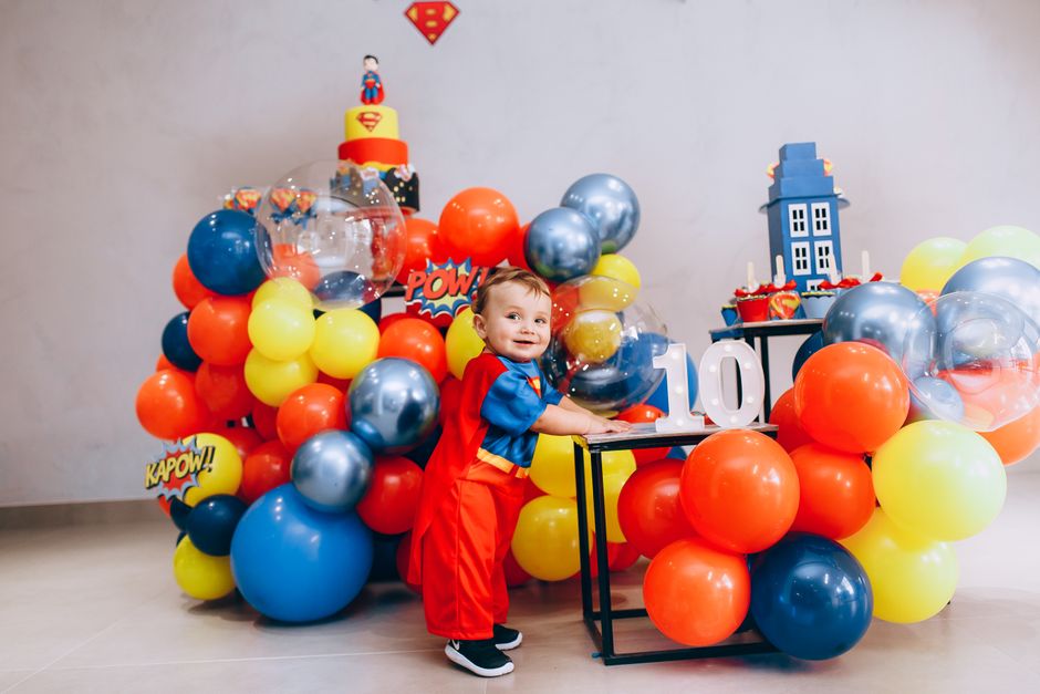 A child in a superhero costume enjoying a colorful birthday celebration indoors with balloons.