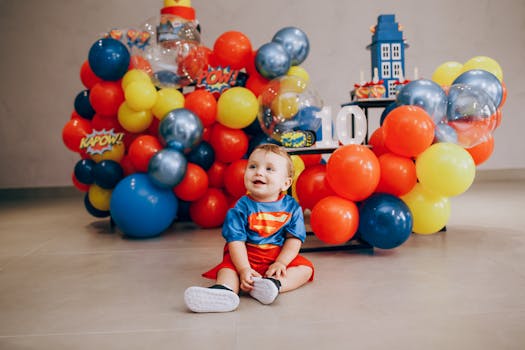 A cute baby dressed as Superman, sitting among vibrant balloons and decorations, radiating joy.